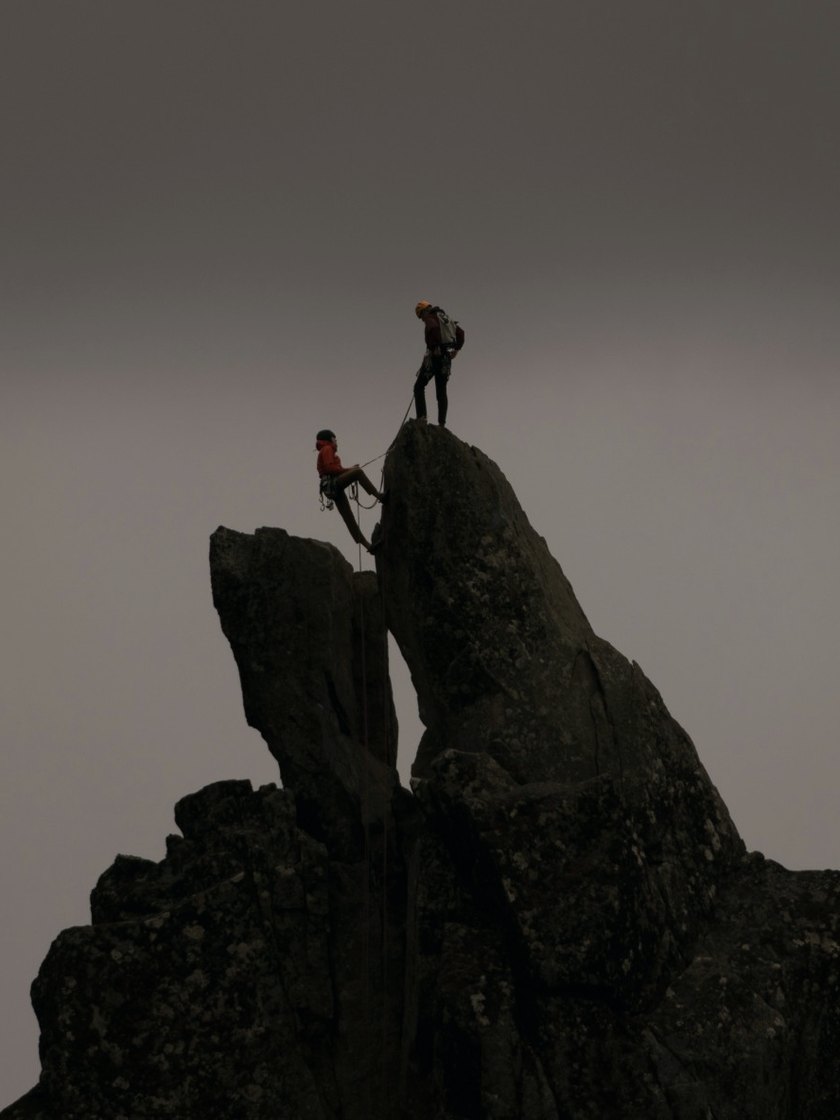 Two climbers on a rock spire, one belaying the other up on a rope